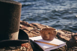 Showing the Kupilka Moomin Pappa in Brown - Classic Cup 210ml by the waterside with top hat and note pad.