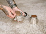 A close-up of assembling a silver aluminium 4 cup moka pot, placing a cafe concetto filter disc on top, with the pot's base set beside it on a textured marble counter top.