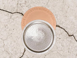 A moka pot lid and metal filter assembly placed on a cracked light beige surface. The lid is peach-colored with the text 'CAFE CONCETTO MOKA SUPERFINE' printed around the edge.