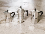 Three moka pots of different sizes arranged on a marble counter top. Bialetti 3 cup and Bialette 4 cup, plus an Alessi stove top coffee maker
