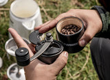 Petromax Hand Coffee Grinder in mans hands opened with coffee beans inside. Coffee set-up in background on grass