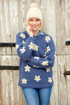 Pachamama Flower Power Sweater in Denim colour with white and yellow daisy designs on blond model wearing a white bobble hat while picking her teeth.