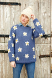 Pachamama Flower Power Sweater in Denim colour with white and yellow daisy designs on blond model wearing a white bobble hat while scratching her head.