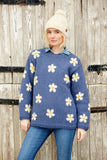 Pachamama Flower Power Sweater in Denim colour with white and yellow daisy designs on blond model wearing a white bobble hat while sticking her leg out.