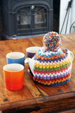 A teapot covered with a Pachamama Yacatan Tea Cosy, on a table with mugs, in front of a stove
